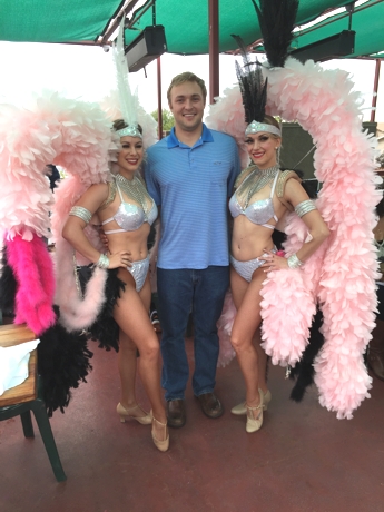 Two women with pink feather headdresses posed with a man in blue shirt.